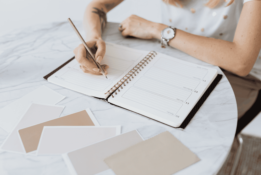 Person writing in a planner at a marble table surrounded by neutral color swatches, symbolizing goal-setting and organization for managing anxiety, overcoming burnout, and achieving New Year resolutions.