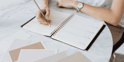 Person writing in a planner at a marble table surrounded by neutral color swatches, symbolizing goal-setting and organization for managing anxiety, overcoming burnout, and achieving New Year resolutions.