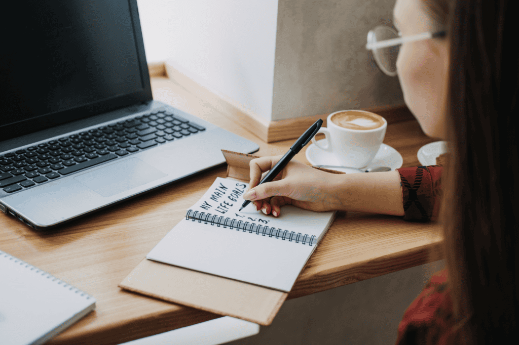 Person planning life goals in a notebook while working at a laptop in a cozy setting, illustrating the importance of mindful goal-setting to prevent high achiever burnout.