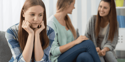 Gen Z girl showing signs of social anxiety symptoms while sitting apart from peers who are laughing and talking.