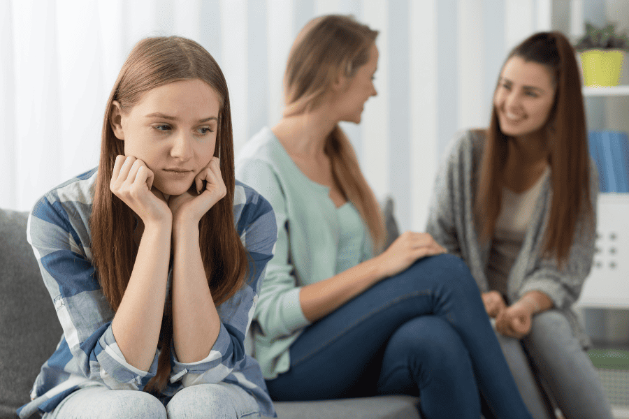 Gen Z girl showing signs of social anxiety symptoms while sitting apart from peers who are laughing and talking.