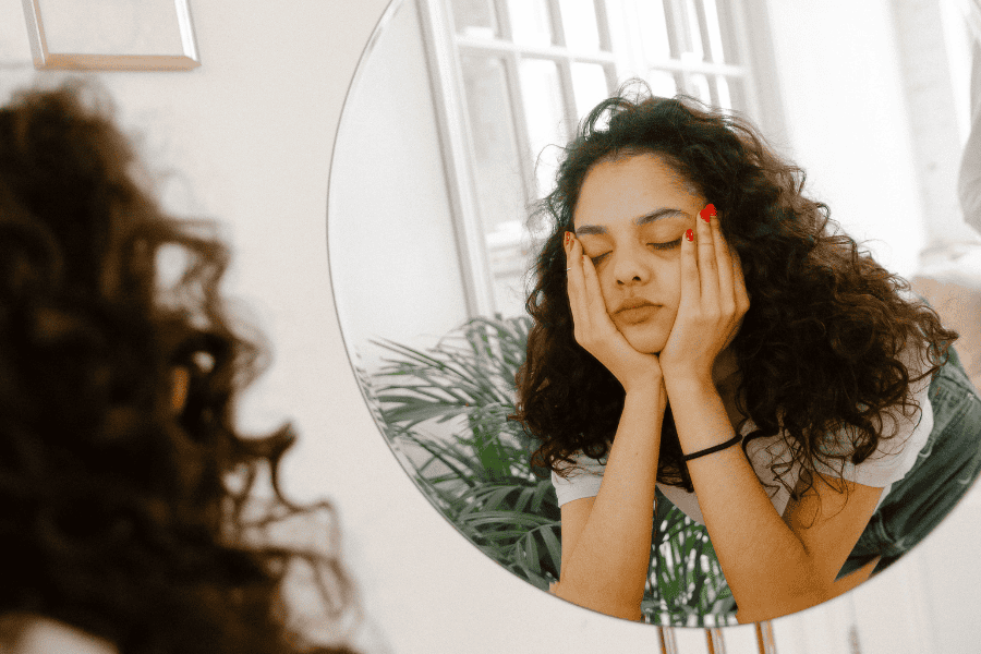 Young woman staring at her reflection with her hands on her face, unsure if she's feeling anxiety or intuition.