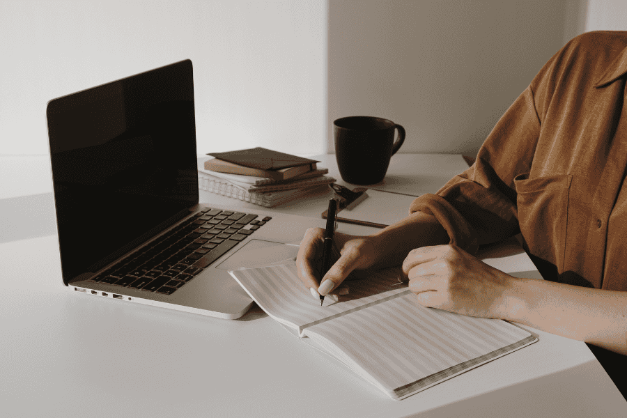 Woman journaling at her desk, reflecting on burnout and toxic hustle culture in a work-from-home setting.