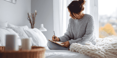 A young woman sitting on her bed and writing in a journal, processing emotions and thinking about why she keeps replaying conversations in her head.