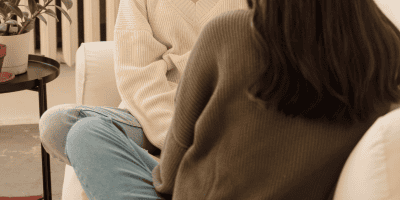 Two friends talking on a couch about personal challenges, reflecting the experience of wondering why you feel responsible for other people’s emotions.