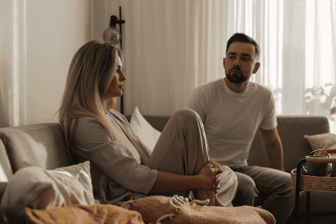 A woman sitting with her knees pulled close while a man looks on with concern, illustrating the tension of feeling responsible for other people’s emotions.